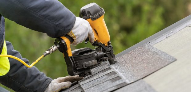 Unrecognizable roofer worker in uniform work wear using air or pneumatic nail gun and installing asphalt or bitumen tile on top of the roof under construction house